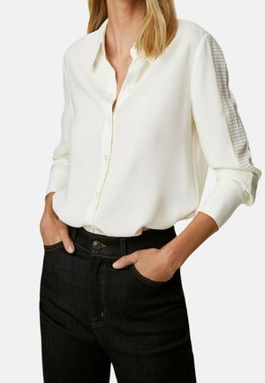 Blusa bianca a maniche lunghe con chiusura a bottoni, caratterizzata da dettagli testurizzati sulle maniche, abbinata a jeans scuri a vita alta.