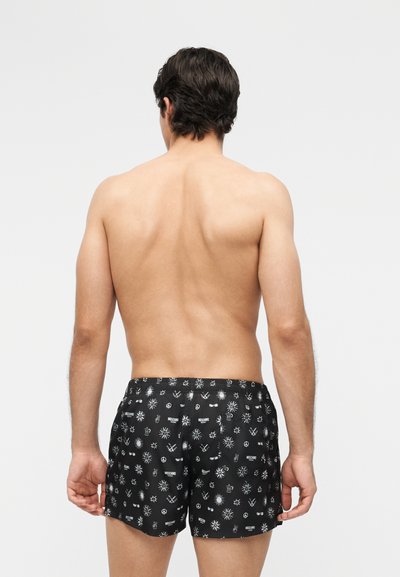 Mann med mørkt hår sett bakfra, iført svarte badeshorts med hvite mønstre av sol, fredstegn og krone på ensfarget bakgrunn.