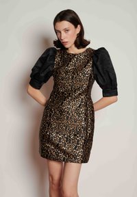 Γυναίκα που φοράει στενό φόρεμα με λεοπάρ μοτίβο και μαύρα φουσκωτά μανίκια μέχρι τον αγκώνα, στεκόμενη μπροστά σε απλό φωτεινό τοίχο.