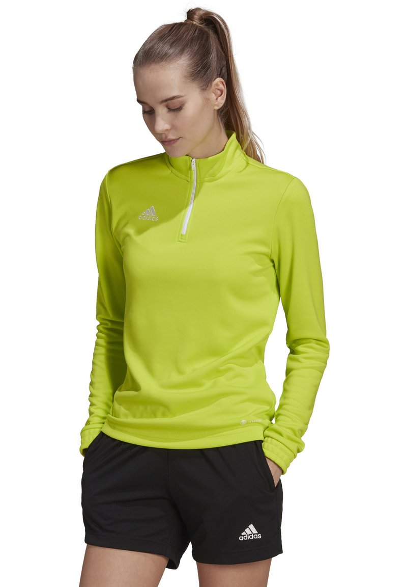 Langärmliges, leuchtend limegrünes Sportoberteil mit einem Viertel-Reißverschluss-Kragen und adidas-Logo. Kombiniert mit schwarzen Shorts. Glattes, leichtes Material.