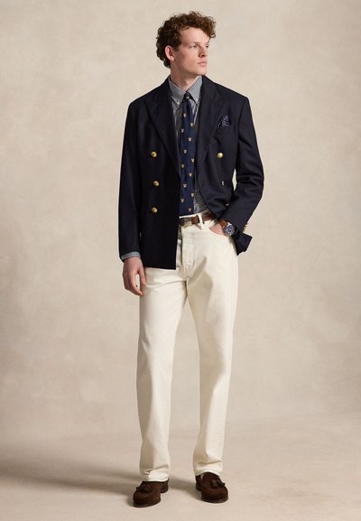 Jeune homme portant un blazer bleu marine croisé, une cravate à motifs, une chemise à carreaux, un pantalon blanc cassé, des chaussures marron à glands et une montre-bracelet, regardant sur le côté.
