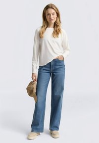 Top bianco a maniche lunghe con texture a coste, abbinato a jeans larghi color azzurro chiaro. La modella è in piedi con scarpe beige e una borsa coordinata.