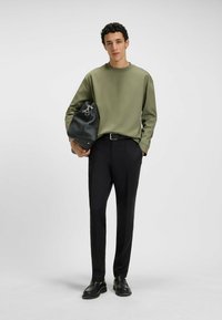 Olive green long-sleeve shirt paired with black trousers; man holding a textured black bag with silver hardware, wearing black shoes.