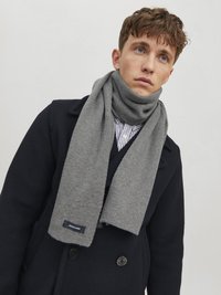 Bufanda gris con textura acanalada, colgada alrededor del cuello, con una etiqueta. Usada con un abrigo azul marino oscuro sobre una camisa a rayas.