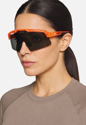 Donna con capelli scuri raccolti che indossa occhiali da sole sportivi oversize con montatura trasparente arancione brillante.