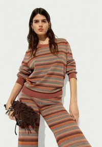 Pull en maille aux rayures rouille, beige et grises, avec des poignets et un ourlet côtelés, associé à un pantalon large assorti. Le mannequin tient un sac marron à franges.