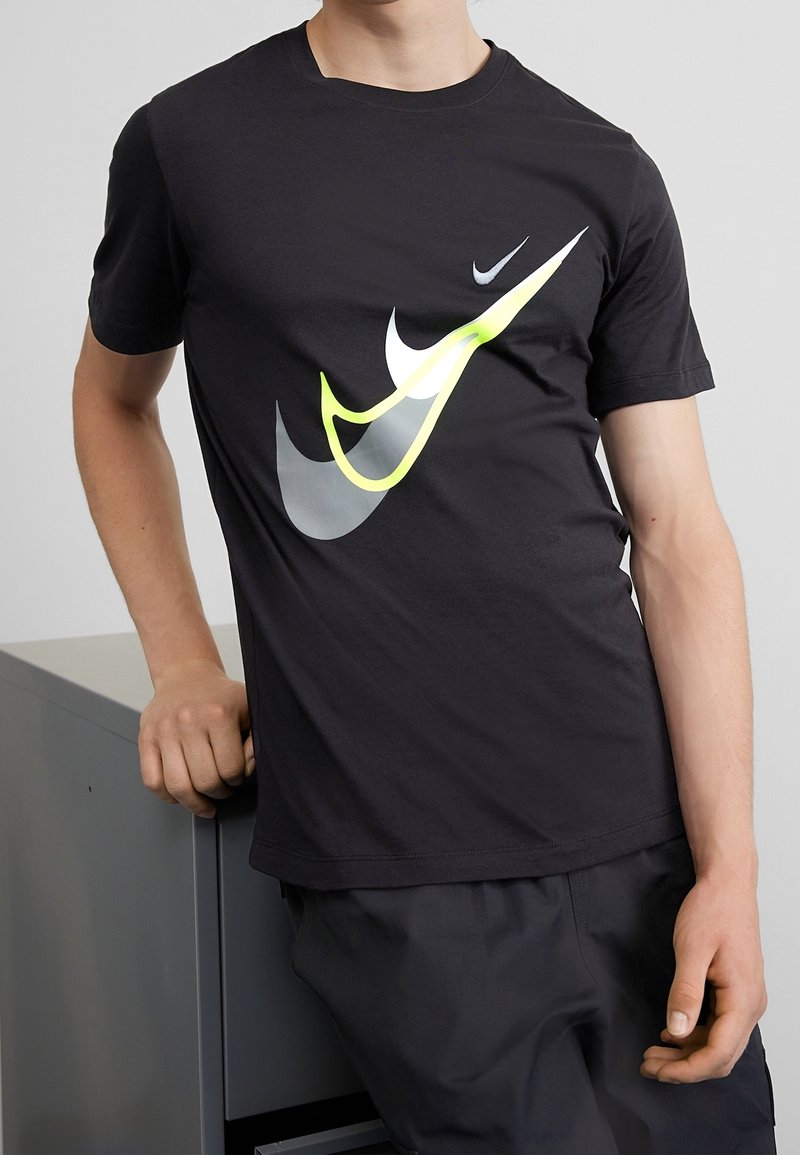 Černé bavlněné tričko s velkými bílými a neonově žlutými logy Nike Swoosh. Krátké rukávy a klasický kulatý výstřih.