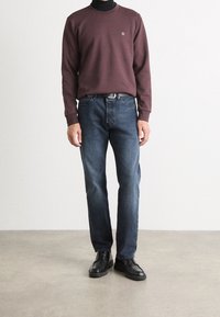 Hombre con un suéter burdeos sobre un cuello de tortuga negro, jeans azul oscuro, cinturón negro y zapatos de cuero negros en un suelo gris con fondo blanco.