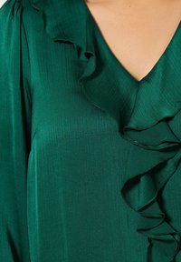 Grüne Bluse mit V-Ausschnitt und strukturierter Stoffqualität, die entlang der Vorderseite und Schultern mit Rüschen gestaltet ist. Glattes, seidiges Erscheinungsbild.