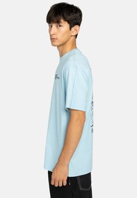 Camiseta de algodón azul claro con mangas cortas, que presenta texto impreso en negro y un diseño gráfico en la parte posterior. Corte recto, ajuste relajado.