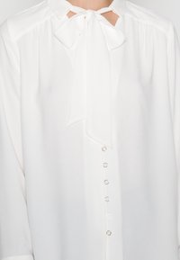 Blusa bianca con colletto, aperture a forma di chiave e fiocco. Presenta una chiusura frontale con bottoni e accenti in metallo argentato, con texture morbida.