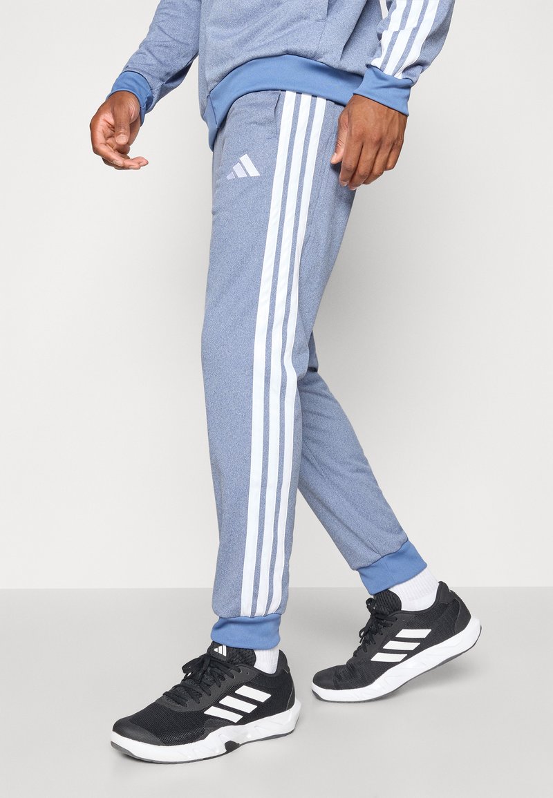 Pantalon de sport bleu clair avec trois rayures blanches sur les côtés, en tissu texturé et avec des poignets élastiques. Assorti à des chaussures de sport noires.
