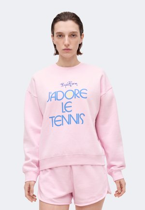 Person wearing light pink sweatshirt and matching shorts with blue text "J'ADORE LE TENNIS" on the front, standing against plain background.