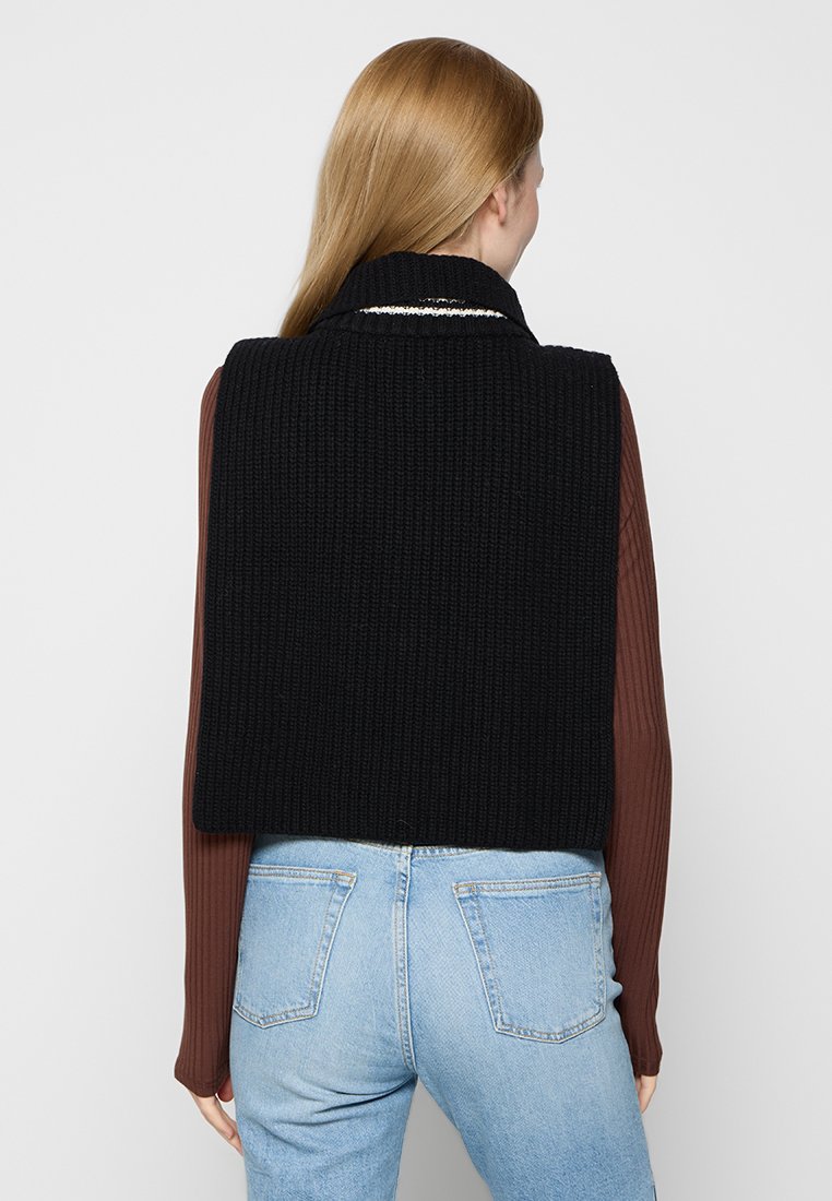 Vrouw met lang blond haar, gekleed in een zwart gebreid mouwloos vest over een bruin geribbeld longsleeve shirt en lichtblauwe jeans, staand met haar rug naar de kijker.
