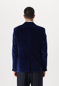 Marineblauwe fluwelen blazer met een enkele split aan de achterkant. Opvallende kenmerken zijn een klassieke kraag en decoratieve knoopaccenten op de mouwen.