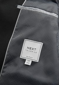 Interior de chaqueta de traje mostrando tela negra con forro estampado geométrico gris y una etiqueta blanca que dice "NEXT Tailoring Slim Fit 40R Made in Vietnam."