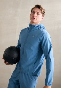 Blå atletisk jakke med lynlås foran og hætte, prydet med et sølvfarvet Nike-logo. Modellen holder en sort tekstureret basketball.