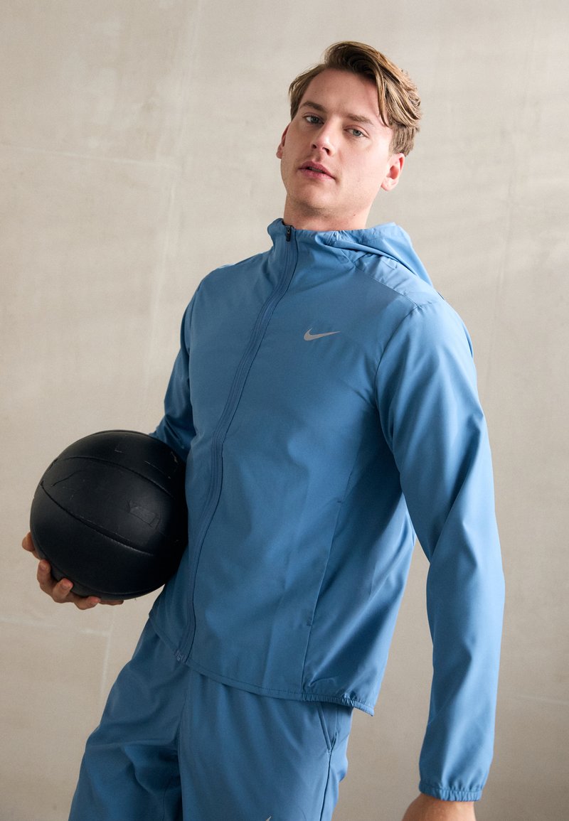 Blå atletisk jakke med lynlås foran og hætte, prydet med et sølvfarvet Nike-logo. Modellen holder en sort tekstureret basketball.