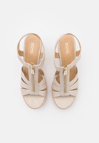 Sandalias de tacón alto beige con parte superior de tela, diseño de punta abierta y detalle de tiras cruzadas. Cuenta con una cremallera lateral y una suela texturizada.