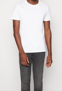 Man som bär en enkel vit t-shirt med kort ärm och bleka svarta jeans, står mot en enkel ljus bakgrund, med överkropp och armar synliga.