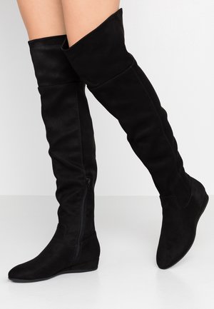 Jambes portant des bottes plates noires au-dessus des genoux avec une fermeture éclair sur le côté, se tenant sur un sol blanc contre un mur blanc.
