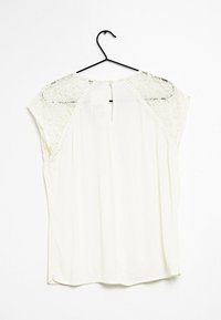 Blouse blanche avec manches courtes en dentelle, dos en forme de goutte avec fermeture à bouton, et tissu lisse et fluide.