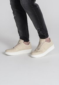 Beige semišové tenisky s textúrovaným zvrškom, s bielou gumovou podrážkou a systémom šnurovania. Nosí sa s roztrhnutými čiernymi džínsm.