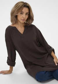Tunic top marrone scuro con scollo a V, maniche a tre quarti e risvolti. Tessuto morbido con una vestibilità ampia, abbinato a jeans denim blu.