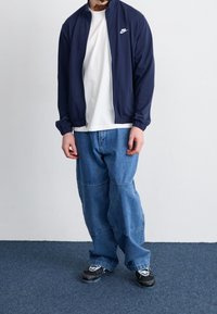Granatowa kurtka zapinana na zamek, biały nadruk, w zestawie z białym T-shirtem, luźne niebieskie spodnie dżinsowe oraz czarne buty sportowe z białymi elementami.