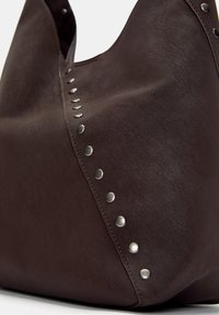 Sac à main en cuir marron avec une texture lisse et des clous métalliques sur un côté. Dispose d'une ouverture sur le dessus et d'une forme arrondie.