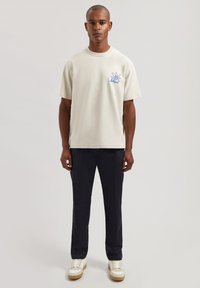 Camiseta beige de algodón con mangas cortas y un diseño gráfico azul en el pecho izquierdo, combinada con pantalones azul marino y zapatillas blancas.