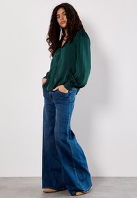 Blusa verde a righe con bottoni e maniche a sbuffo abbinata a jeans denim blu a gamba larga e scarpe beige. Design di un outfit semplice e casual.