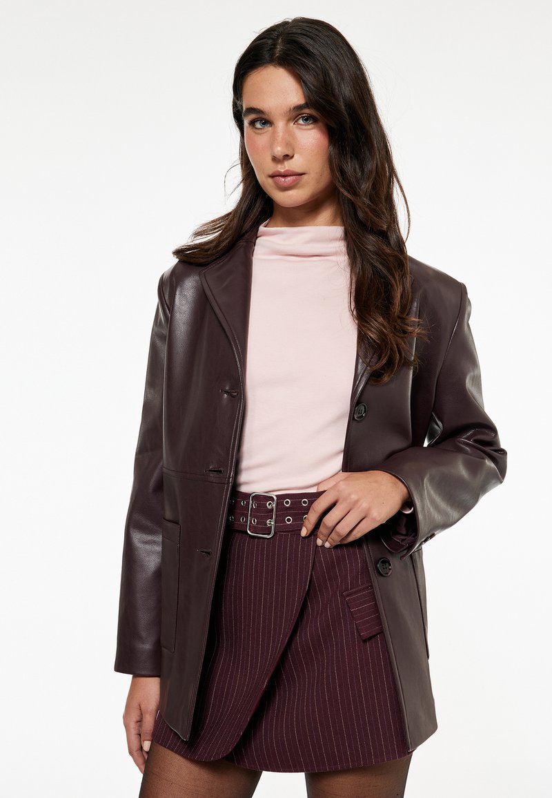 Blazer de cuero burdeos con cuello con muesca, cierre de un botón y bolsillos frontales. Combinado con una blusa rosa claro y una falda a rayas.