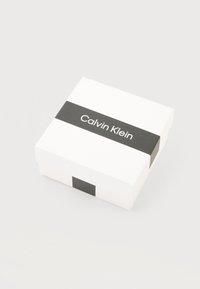 Άσπρο τετράγωνο κουτί με μαύρη οριζόντια λωρίδα που εμφανίζει το λογότυπο "Calvin Klein" στο κέντρο, σε απλό ανοιχτό φόντο.