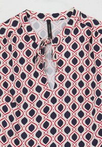 Blusa bianca con un motivo geometrico ripetuto in rosso e blu navy, caratterizzata da un collo a spacco con lacci e una texture di tessuto leggero.