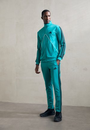 Mann iført en teal Adidas treningsdress med svarte striper og svarte sneakers, som står mot en ensfarget lys grå vegg.