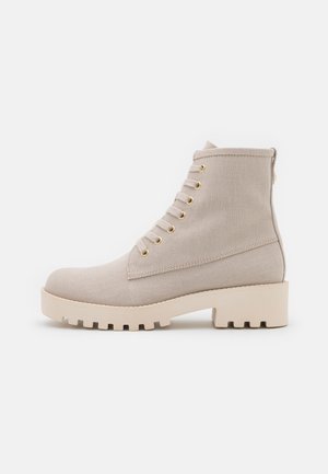 Bottine beige montant à la cheville avec lacets, semelle épaisse à crampons, œillets dorés et tige en tissu texturé, vue de côté sur fond blanc.
