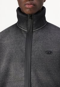 Detailní záběr na osobu oblečenou v tmavě šedé mikině na zip s vysokým límcem a malým černým logem na hrudi.