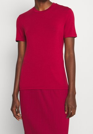 Osoba ubrana w czerwoną, krótkim rękawem i okrągłym dekoltem koszulkę, włożoną w pasującą, plisowaną czerwoną spódnicę, stojąca z ramionami luźno opuszczonymi wzdłuż ciała.