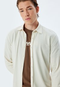 Camisa texturizada de color claro con botones en la parte delantera, con cuello y mangas largas, superpuesta sobre una camiseta marrón con estampado de logo.
