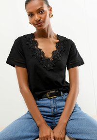 Melna īsām piedurknēm topa ar dziļu V veida kakla izgriezumu, kas rotāts ar ziedu mežģīņu izšuvumu gar kakla līniju un teksturētu audumu. Valkāts ar augsto vidukli džinsiem.