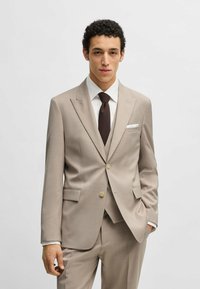Abito beige chiaro con rever a punte, abbinato a una camicia bianca e una cravatta marrone scuro. Presenta bottoni dorati e una tasca sul petto.