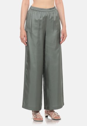 Donna che indossa pantaloni grigi in raso a vita alta e gamba larga e sandali beige con tacco e cinturini, in piedi davanti a uno sfondo bianco semplice.