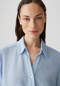 Camicia button-up azzurra chiara realizzata in tessuto testurizzato, con colletto e morbide pieghe sulla superficie. Dettaglio con un solo bottone visibile.