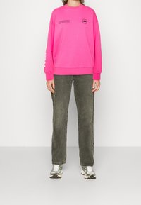 Sudadera rosa brillante con cuello redondo, mangas largas y detalles impresos en negro. Combinada con pantalones de pierna recta en verde oliva y zapatillas plateadas.