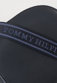 Černá kožená taška se širokým námořnickým popruhem, na kterém je vyšitý nápis "TOMMY HILFIGER" modrou barvou. Hladký povrch s viditelnými švy.