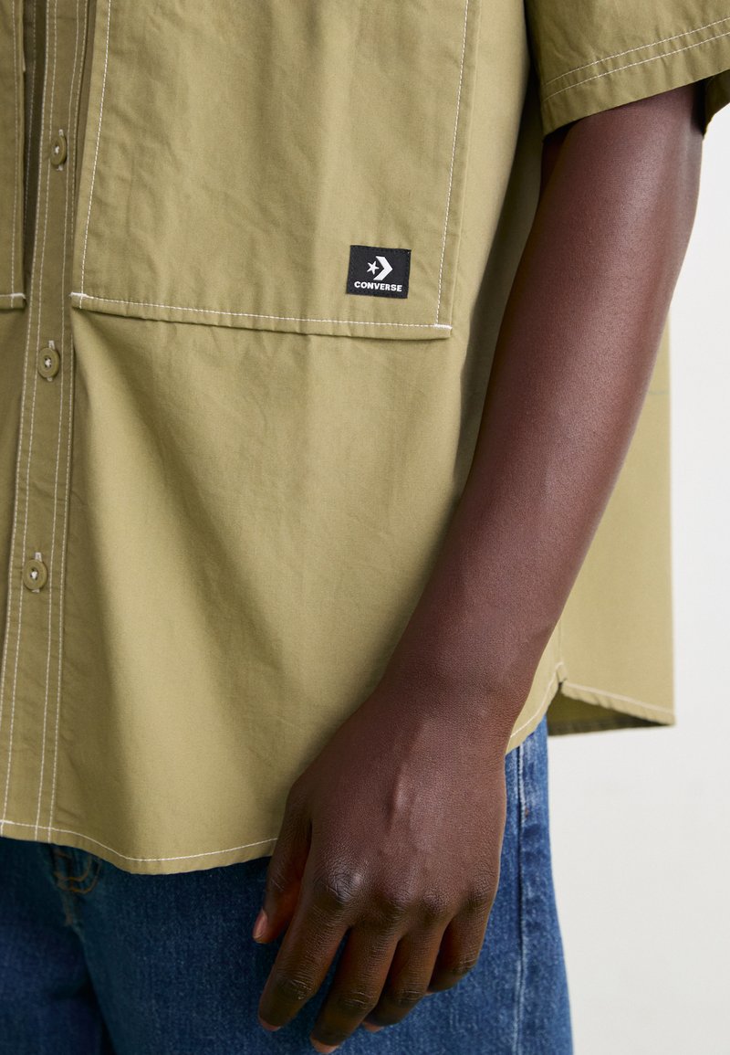 Camisa de manga corta verde oliva con frente abotonado, dos bolsillos tipo parche y costuras blancas; incluye una etiqueta con el logo de Converse en negro.