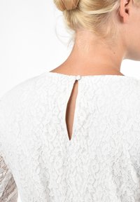 Biała koronkowa bluzka z wycięciem na plecach, zamykana małym guzikiem, z teksturowanym kwiatowym wzorem na całej powierzchni.