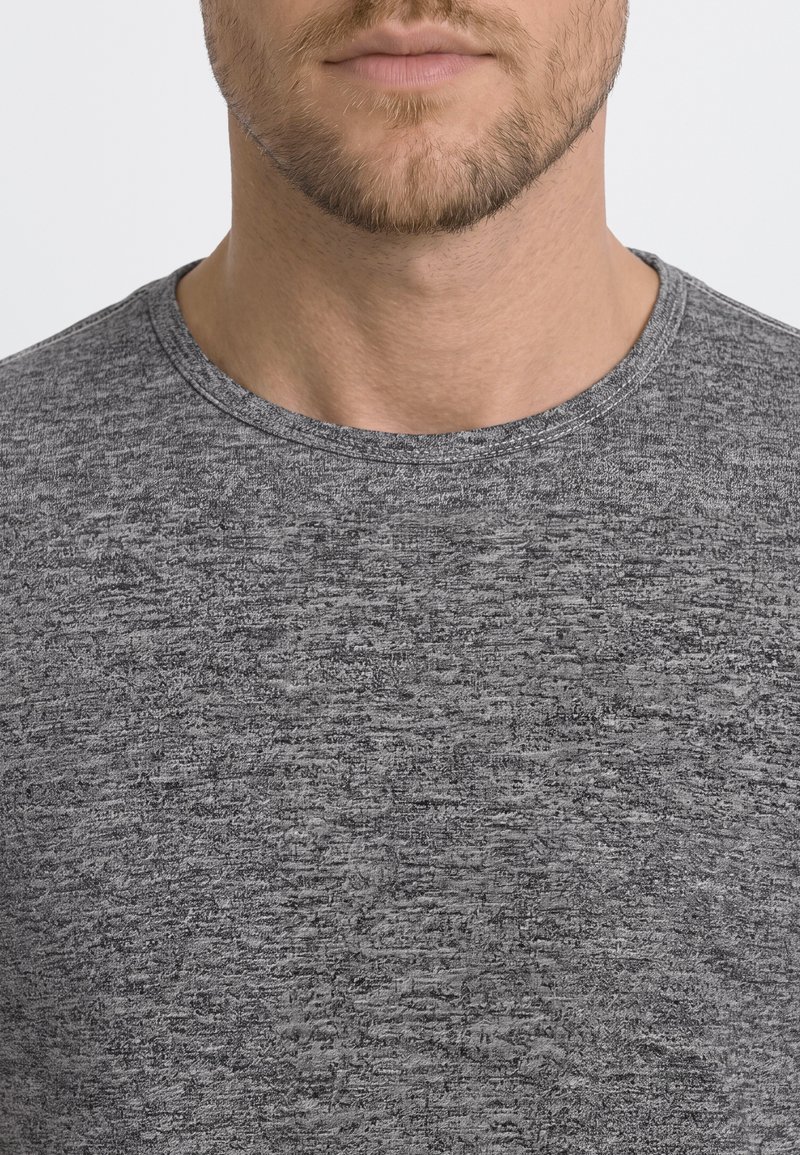 Gros plan sur un homme barbu portant un T-shirt gris chiné à col rond, sur un fond clair uni.