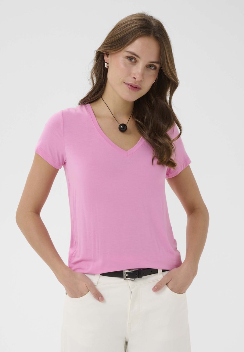 Žena s dlhými hnedými vlasmi má na sebe ružové tričko s véčkovým výstrihom a krátkymi rukávmi, biele nohavice s čiernym opaskom, čierny prívesok na náhrdelníku a strieborné kruhové náušnice.